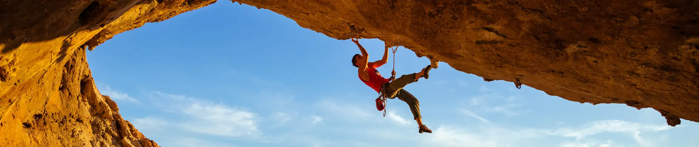 A rock climber hangs from a steep cave ceiling against a clear blue sky background during a sunny day.
