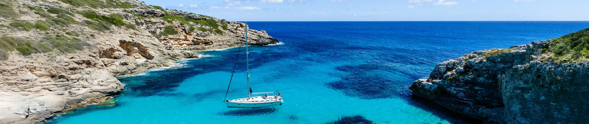 A white sailboat anchors in a beautiful turquoise bay surrounded by rocky cliffs under a blue sky.