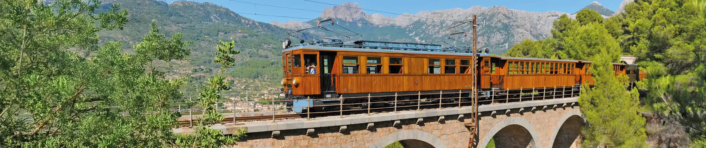 A vintage wooden train crosses a stone arched bridge surrounded by green trees and mountain scenery.