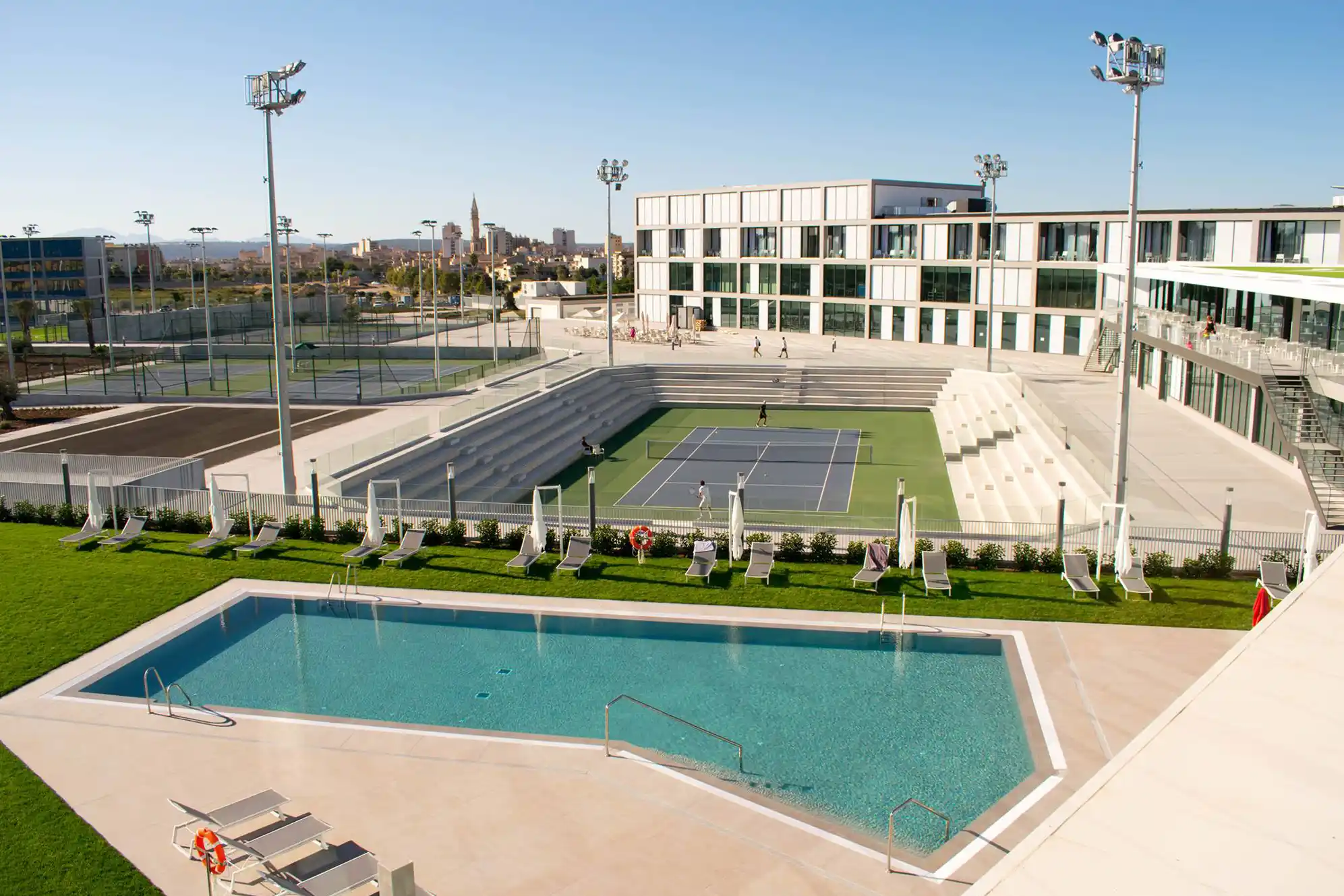 Modern sports center with a swimming pool and tennis courts under a clear blue sky.