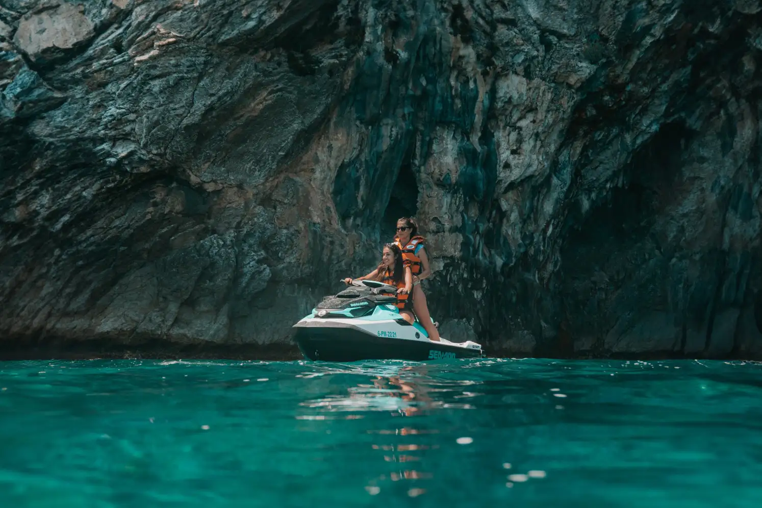 Two people on a jet ski in calm turquoise sea water next to a massive, dark rock formation or sea cave cliff. Two people on a jet ski in calm turquoise sea water next to a massive, dark rock formation or sea cave cliff.