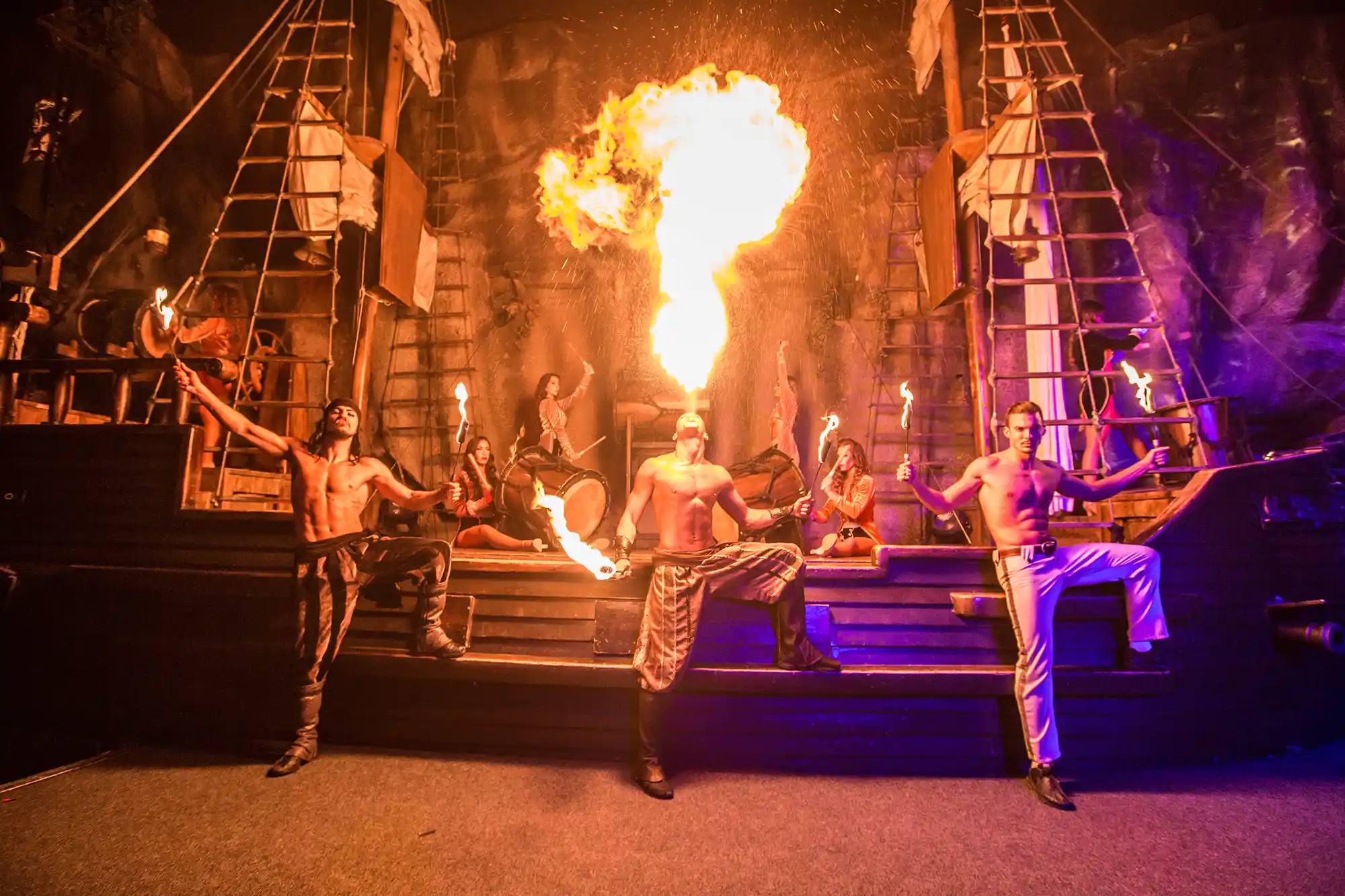 A fire breather performs on a pirate ship stage surrounded by other actors during an impressive show.
