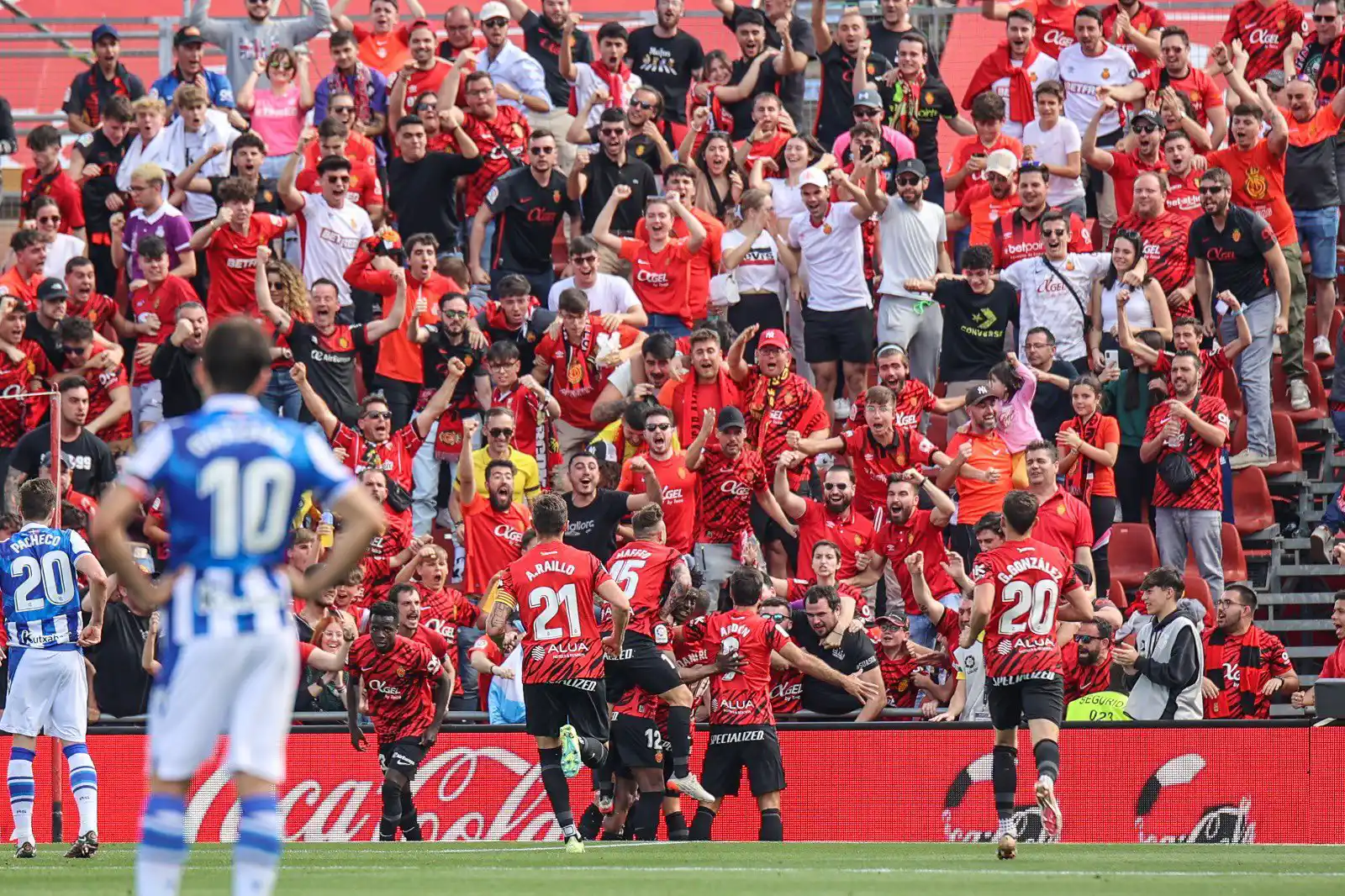 Fußballspieler in Rot feiern ein Tor vor jubelnden Fans, während die Gegner in Blau enttäuscht zusehen.