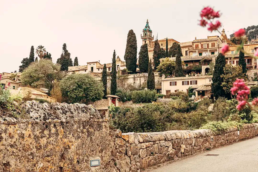 A scenic view of a historic hillside village with stone walls, green trees and a church tower.