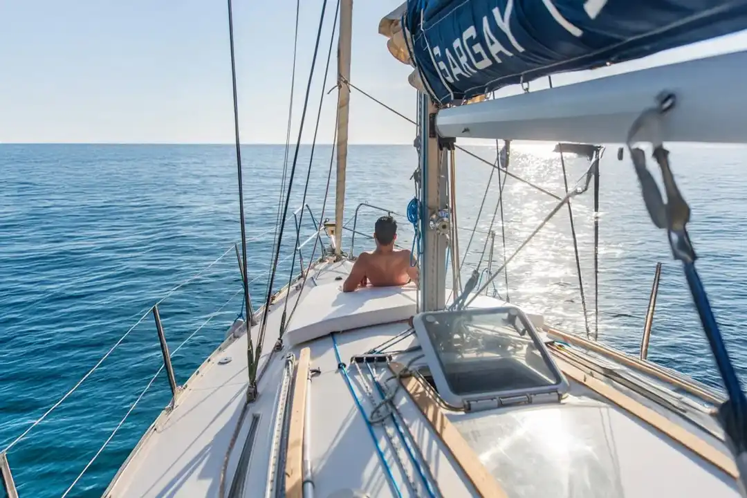 A man relaxes on the deck of a sailboat navigating through calm, deep blue sea water on a sunny day.
