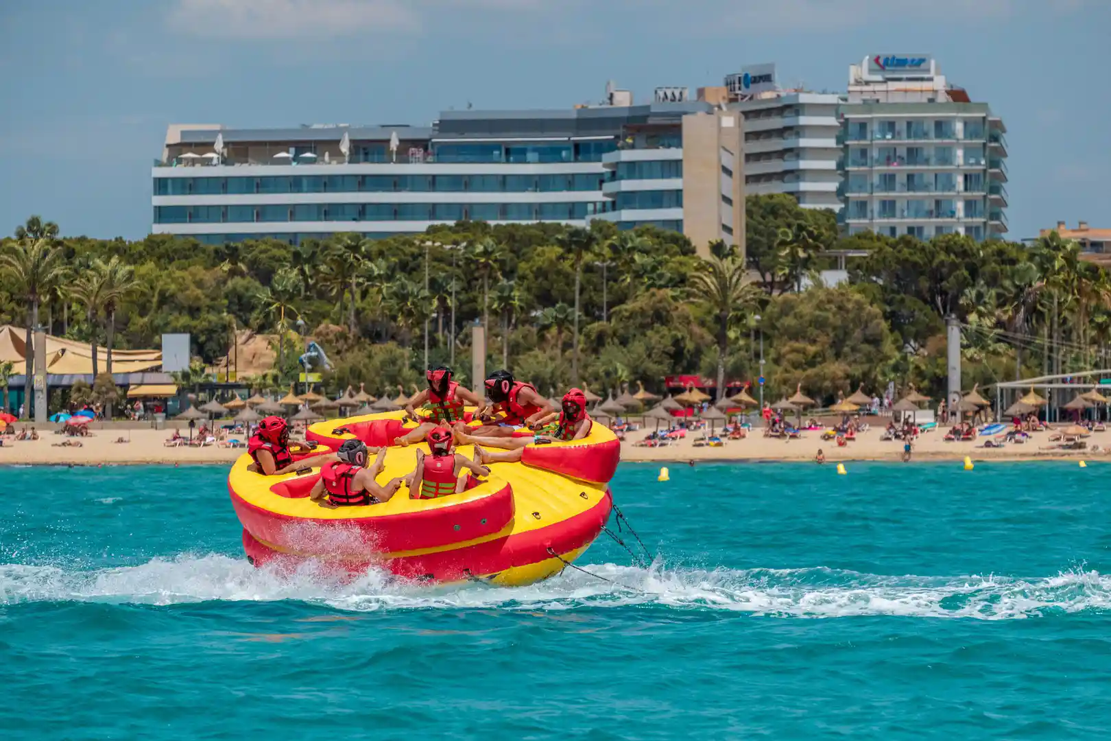 Menschen auf einem runden Bananenboot fahren an einem belebten Sandstrand und Küstenhotels vorbei.