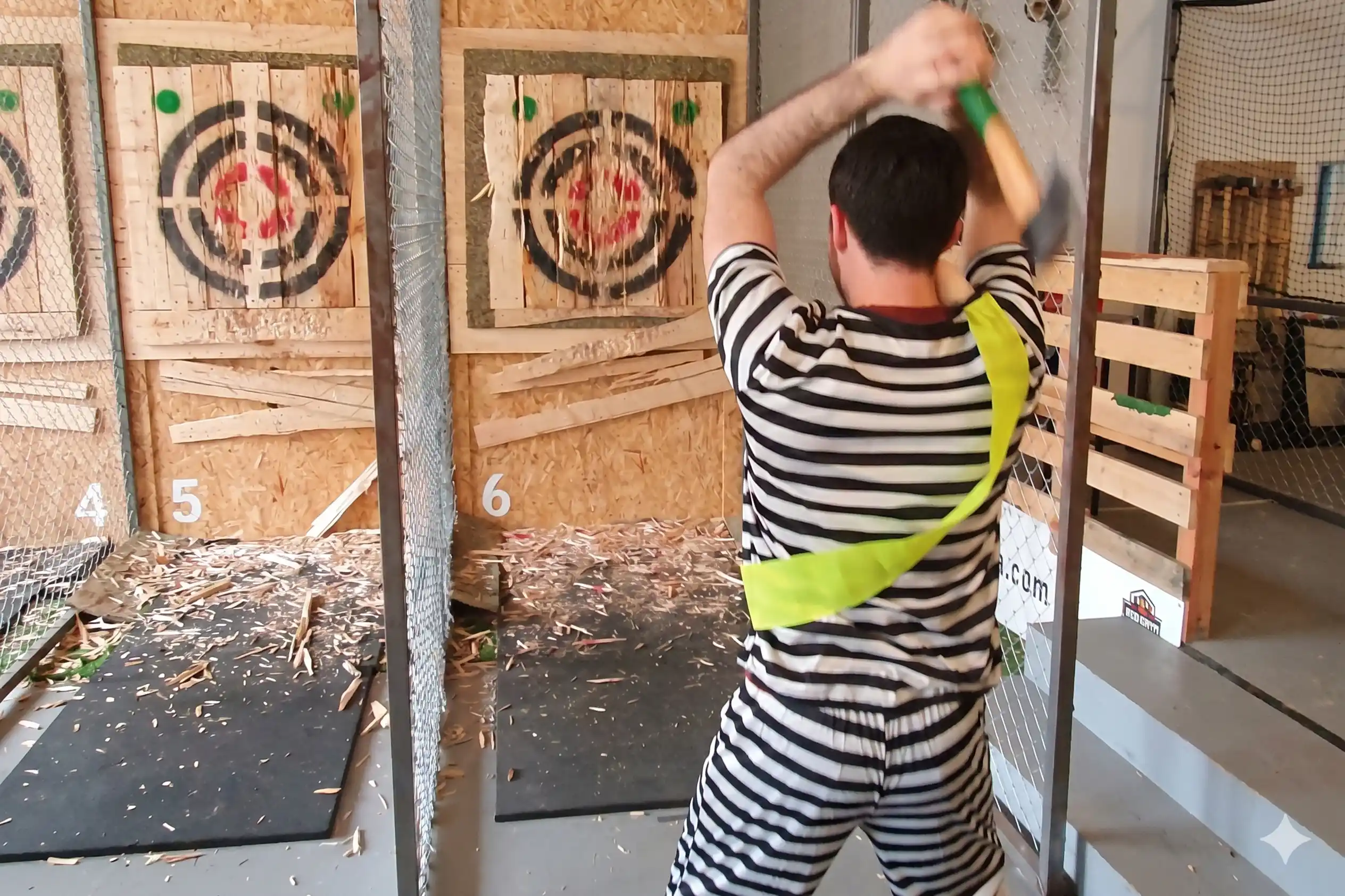 Man in a striped shirt and high-visibility vest raising an axe to throw at a wooden target in an indoor range.