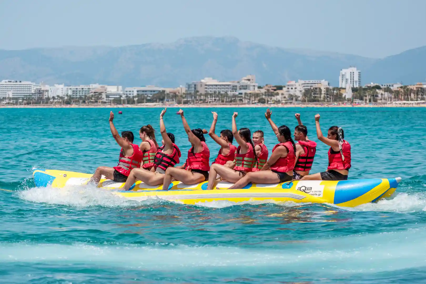 Menschen in roten Schwimmwesten heben die Arme auf einem Bananenboot mit Küstenhotels im Hintergrund.