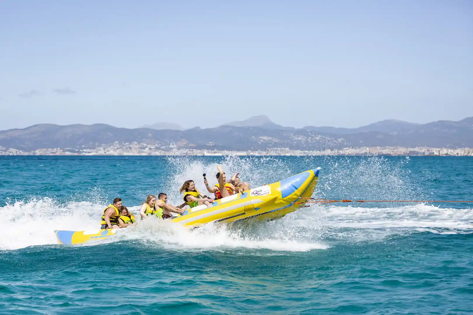 Eine Gruppe von Menschen auf einem gelben Bananenboot springt über die Meereswellen mit Bergen im Hintergrund.