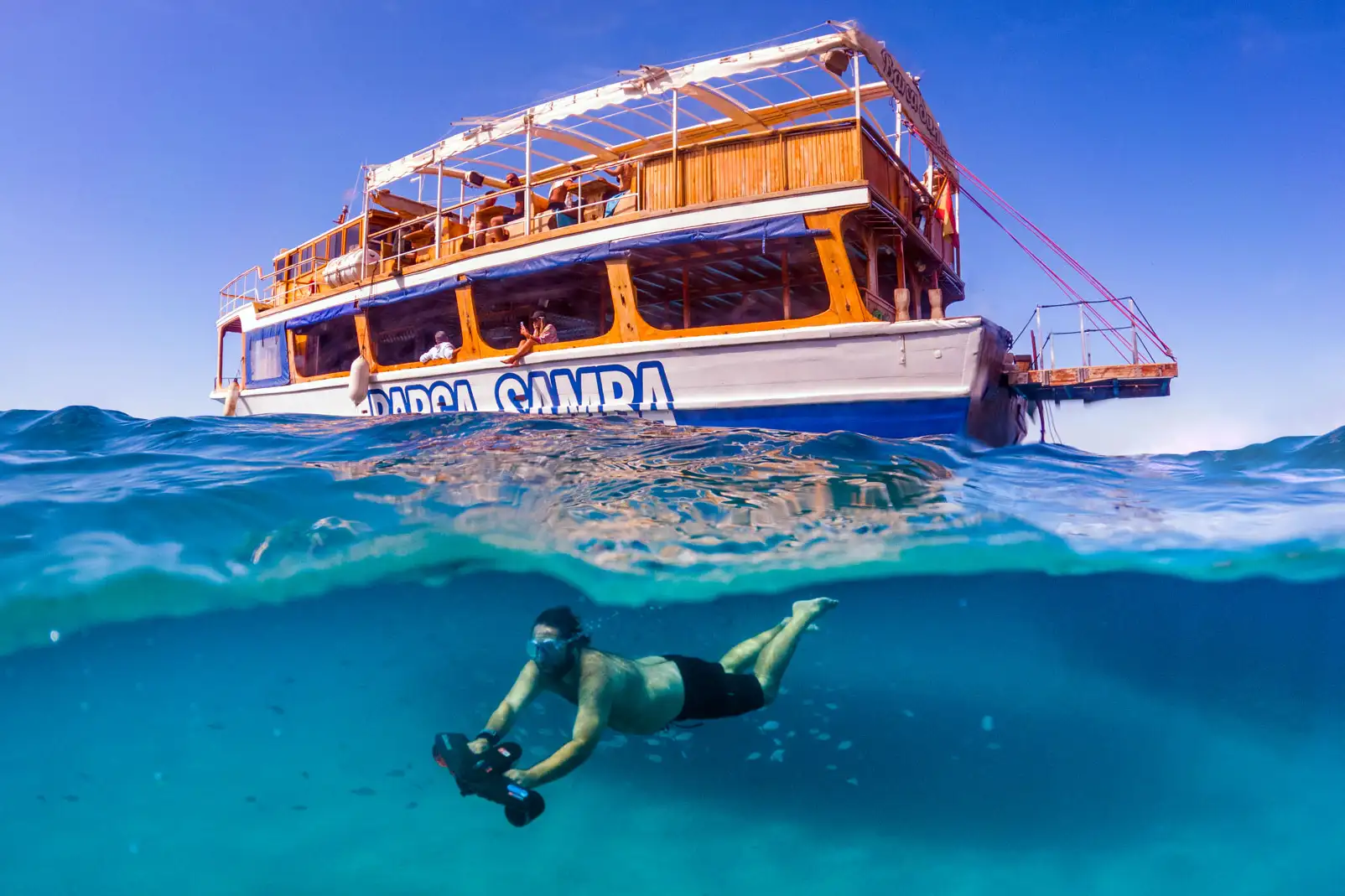 A man using an underwater scooter below the surface while a large tour boat floats above in turquoise water.A man using an underwater scooter below the surface while a large tour boat floats above in turquoise water.