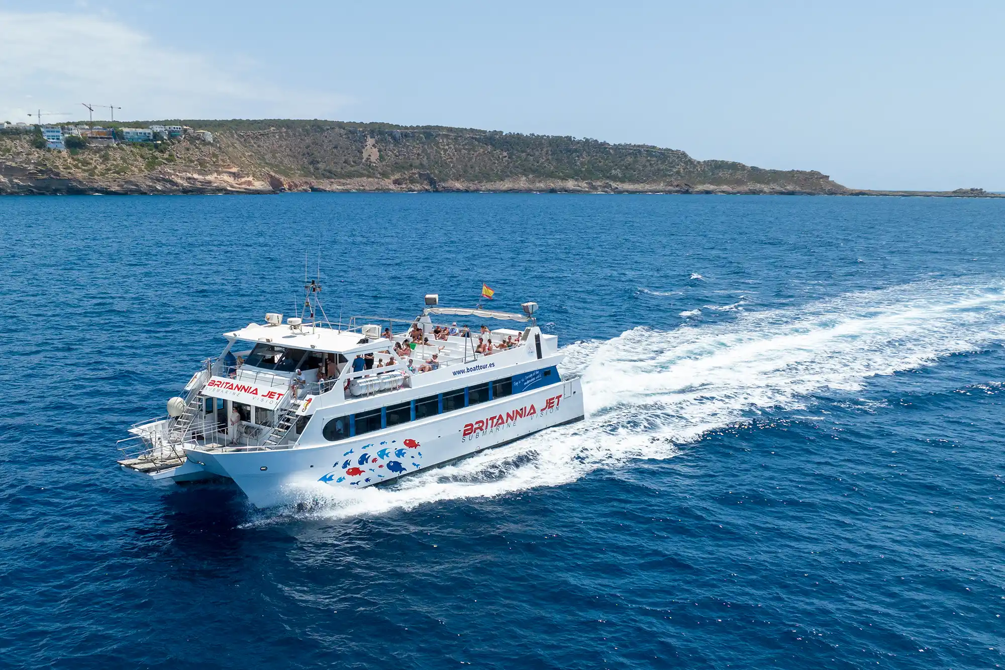 A white catamaran ferry cruises through the deep blue sea, leaving a white wake with a hilly coast behind.