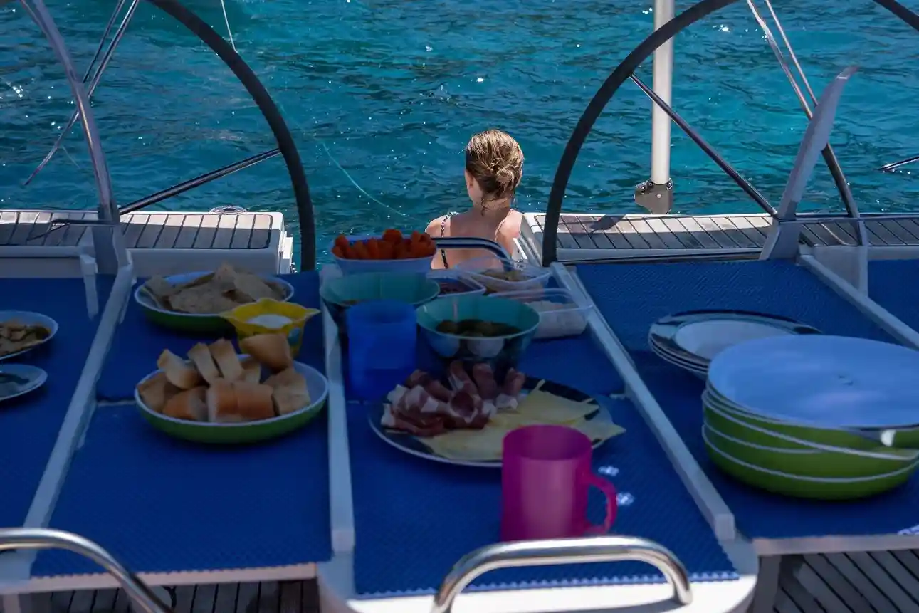 A table with food on a boat deck with a woman looking at the turquoise water in the background.