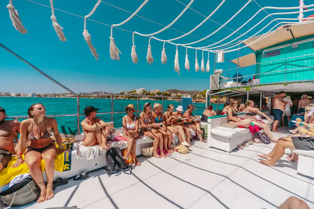 Passengers relax on the deck of a tour boat cruising along a sunny coastline on the blue sea.