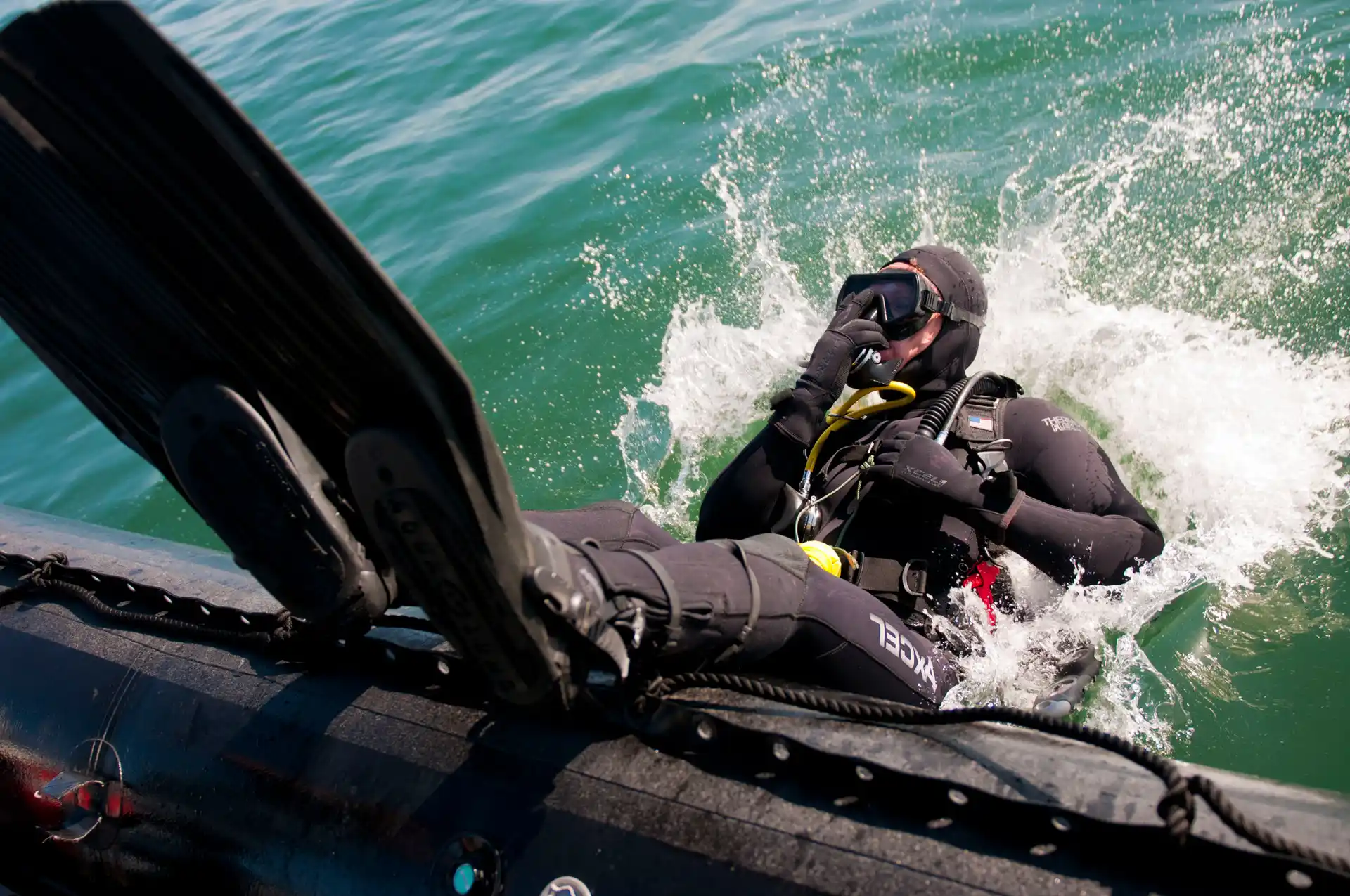 A scuba diver in a black wetsuit falling backward from an inflatable boat into the green sea water.