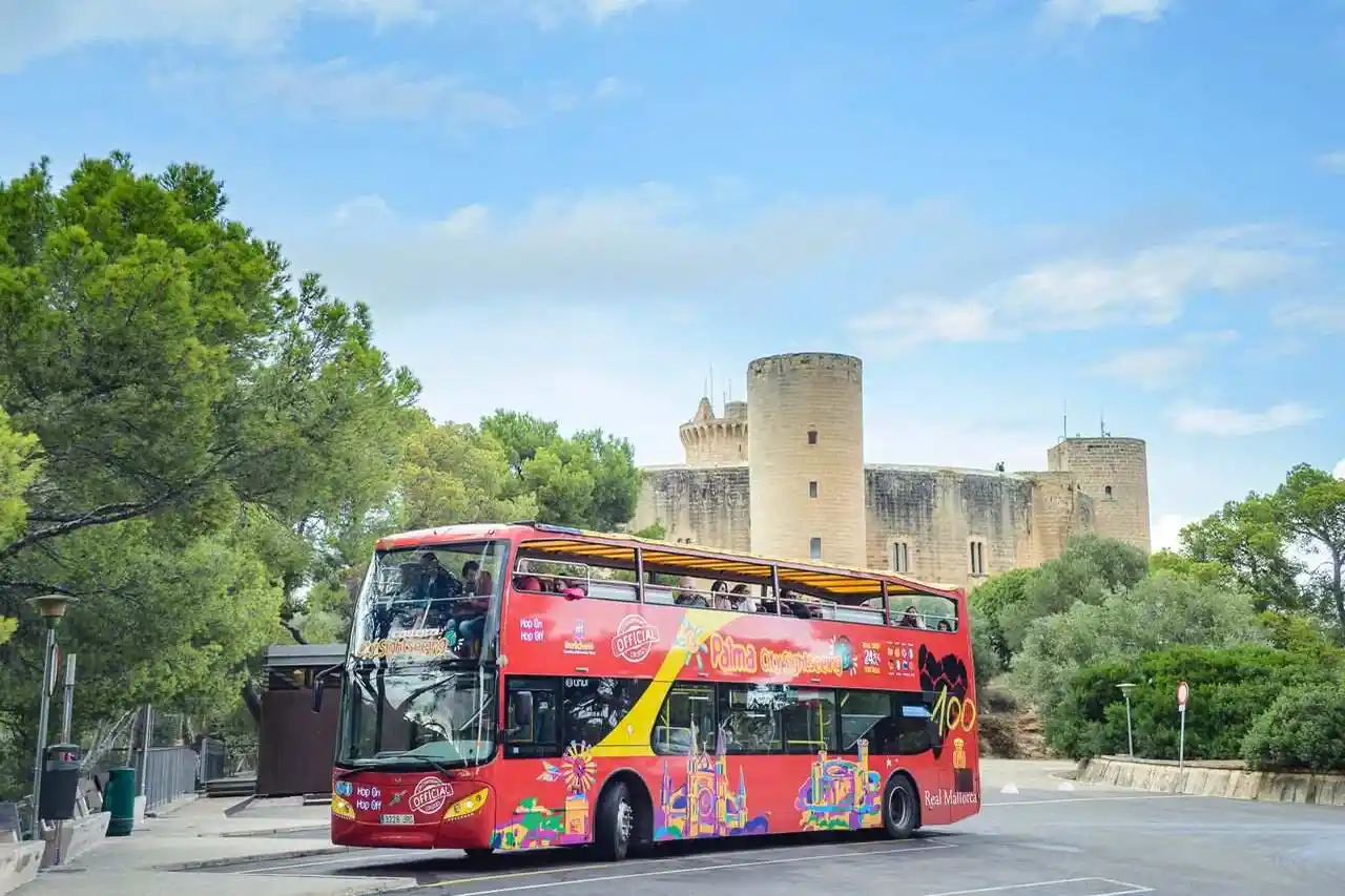 A red double-decker tourist bus drives on a road near a historic stone castle surrounded by green trees under a blue sky.