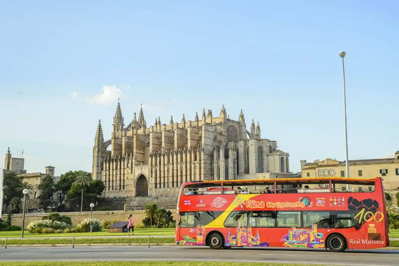 A red double-decker tourist bus drives past a large historic gothic cathedral under a clear blue sky.