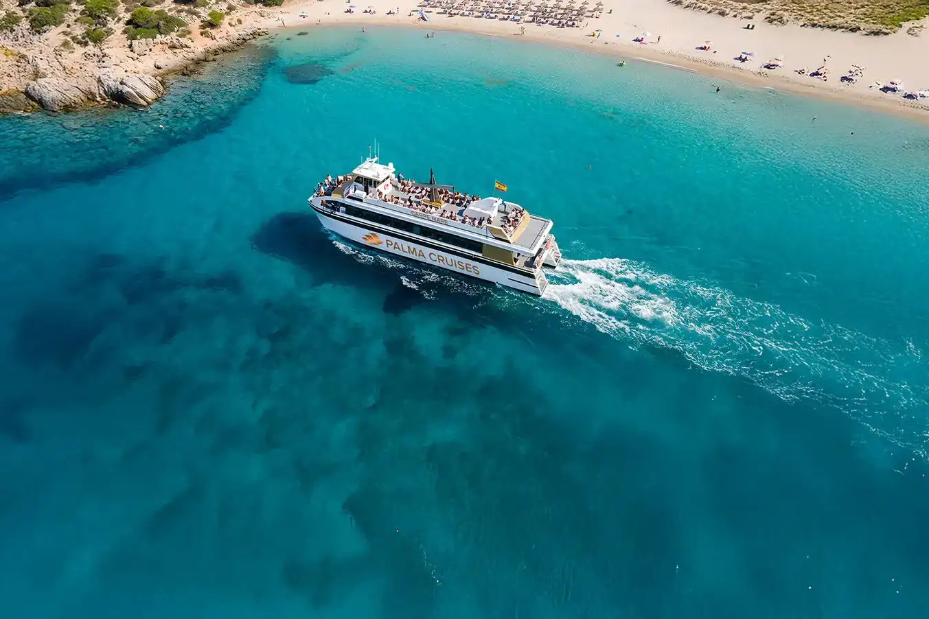 An aerial view of a tourist ferry boat cruising near a sandy beach through clear turquoise coastal water.