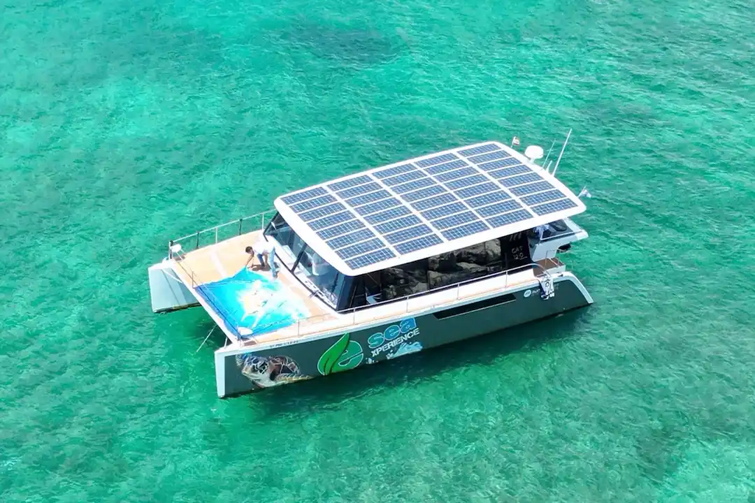Une vue aérienne d'un catamaran moderne équipé de panneaux solaires sur son toit flottant dans une eau turquoise.