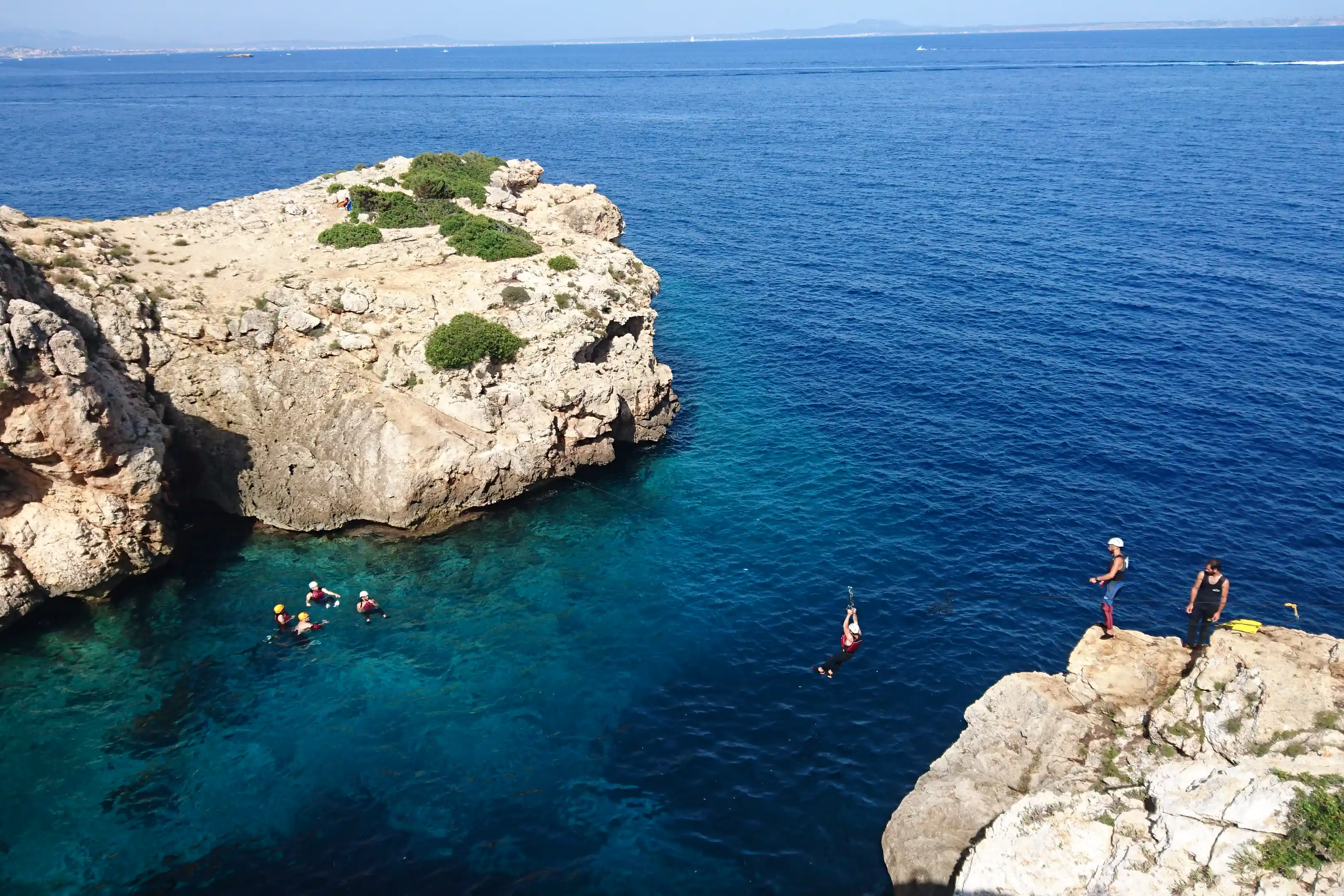 People swimming in a crystal-clear cove while someone ziplines between rocky cliffs above the sea.