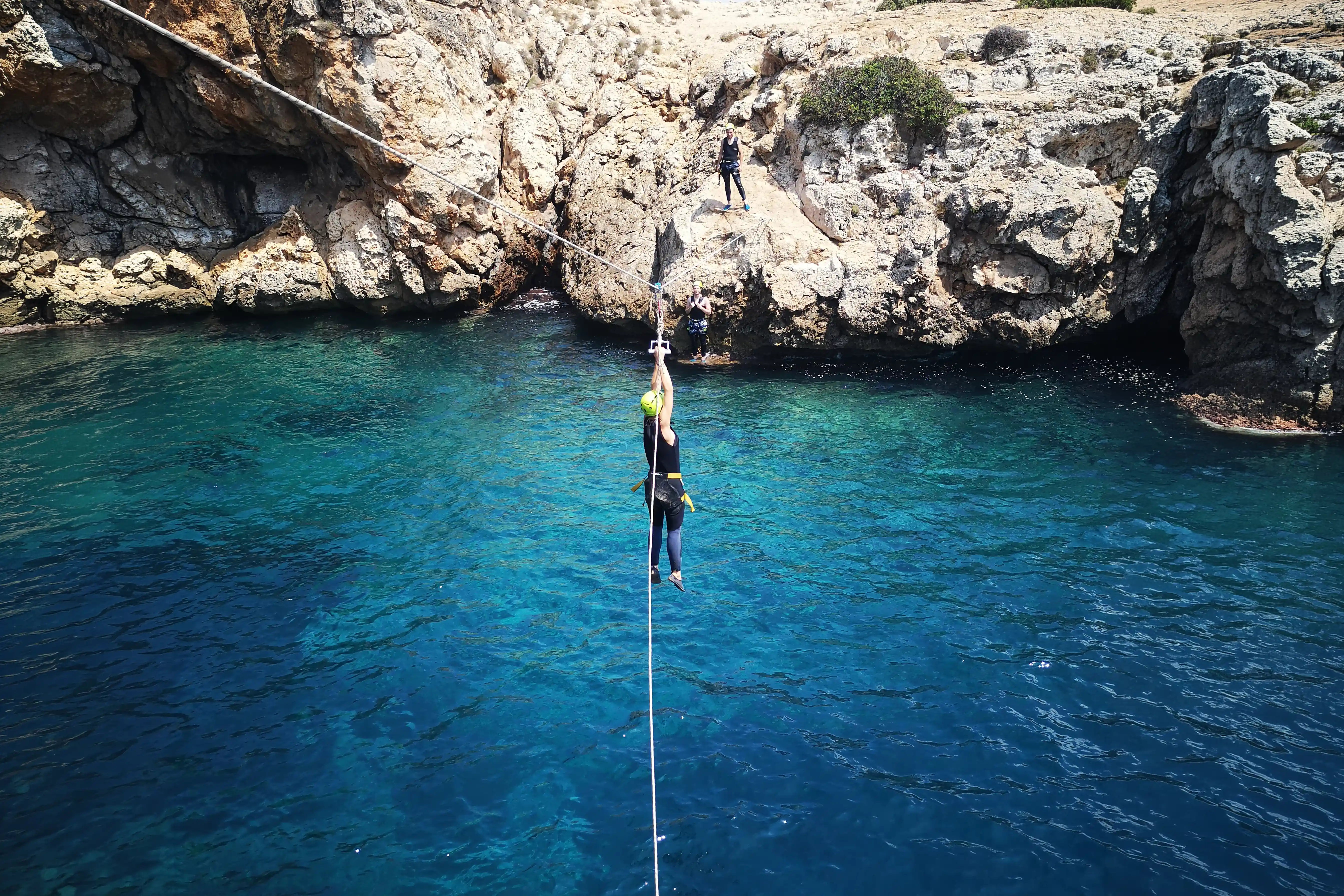 A person hanging from a zip line high above clear, deep blue sea water surrounded by rocky coastal cliffs.