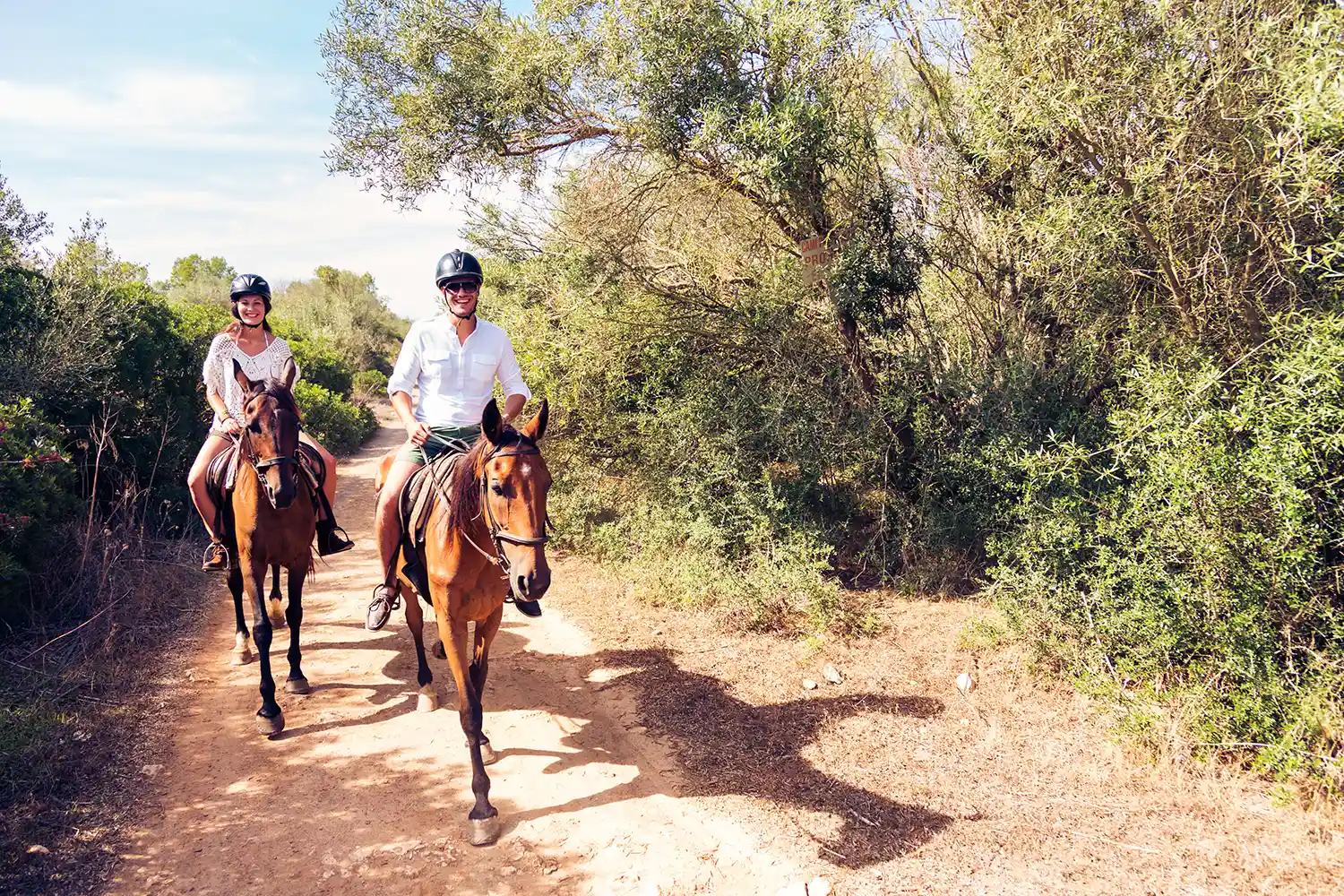 A smiling man and woman wearing helmets ride brown horses along a sunny dirt path surrounded by trees.