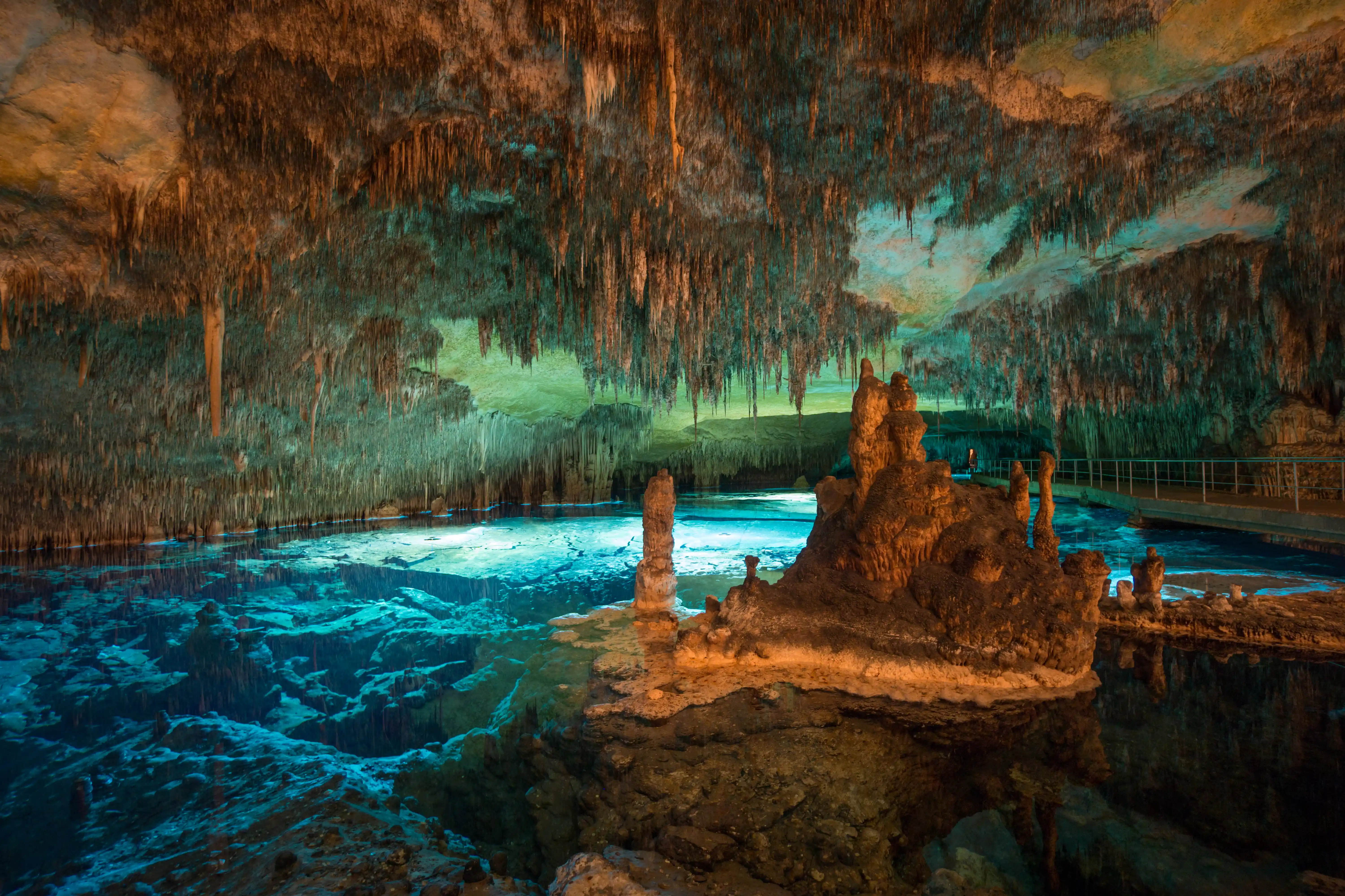 An illuminated blue underground lake inside a large cave filled with rock stalactites and stalagmites.