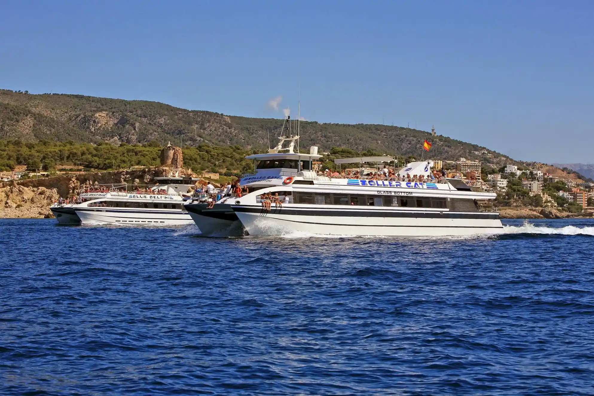 Two large tourist catamaran boats cruise across the deep blue sea along a hilly coastline on a sunny day.
