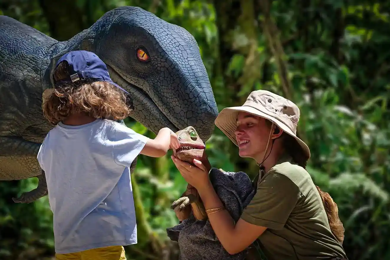 A child and a guide interact with a small dinosaur puppet in front of a large realistic velociraptor.