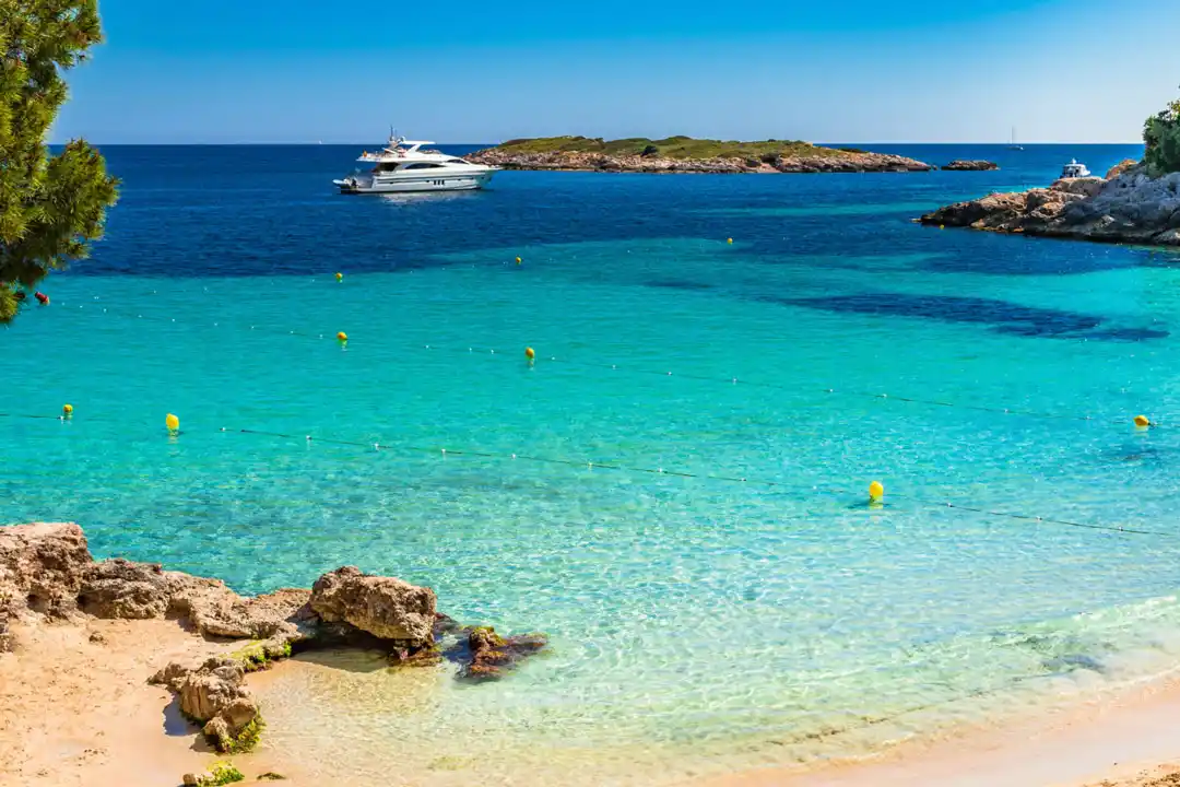 A white yacht anchors in a beautiful turquoise cove with crystal clear water next to a rocky shoreline.