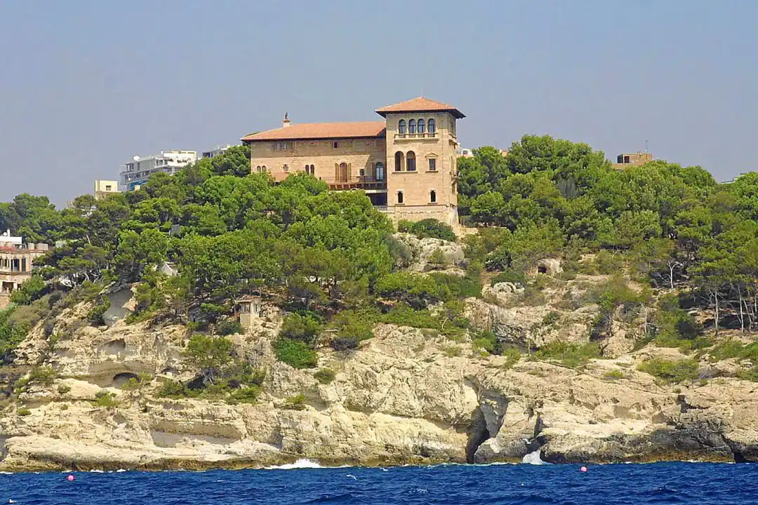 Une villa historique avec une tour s'élève sur une colline verdoyante surplombant une côte rocheuse et la mer bleue.