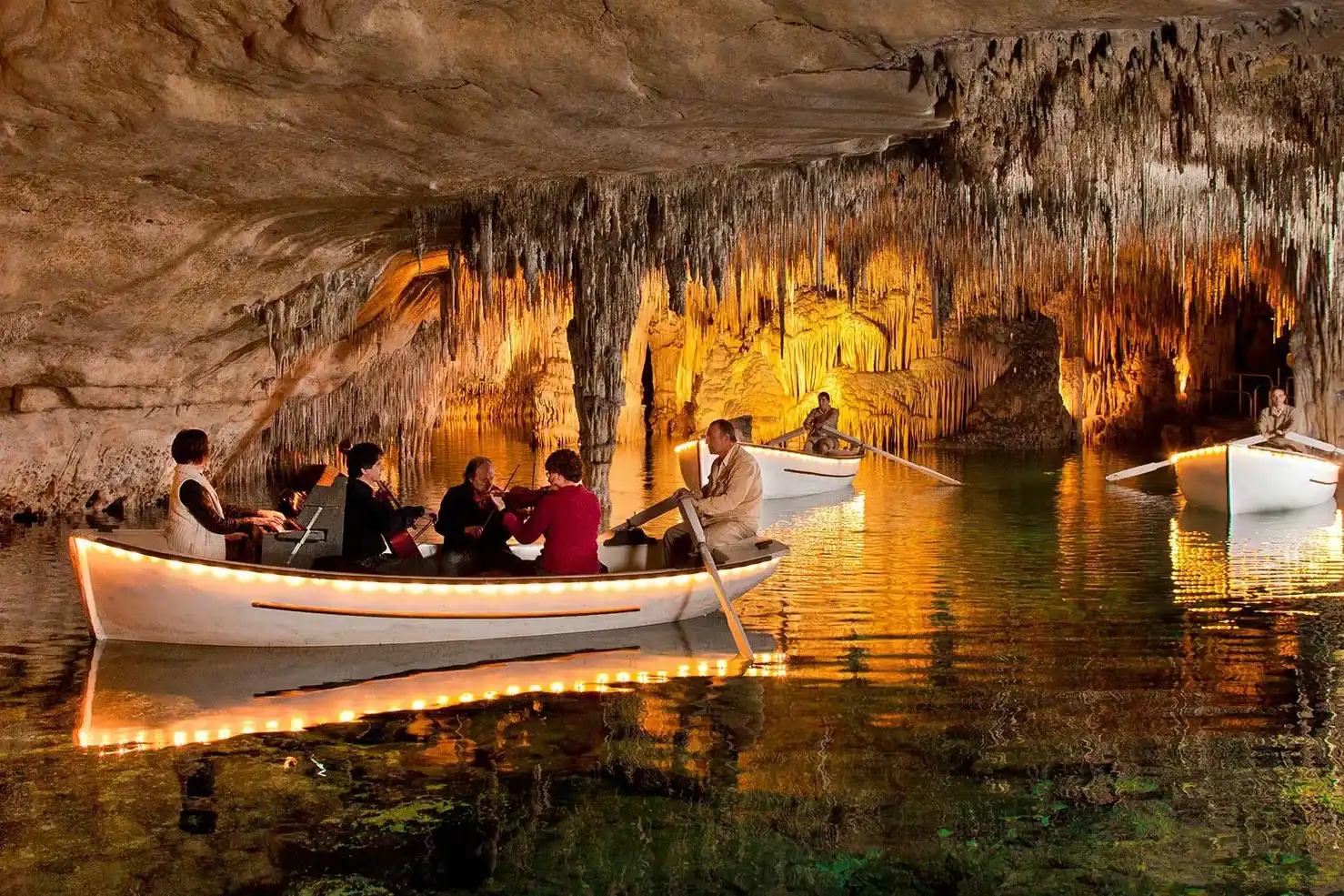 Tourists ride in small boats on an underground lake inside a large illuminated cave with stalactites.