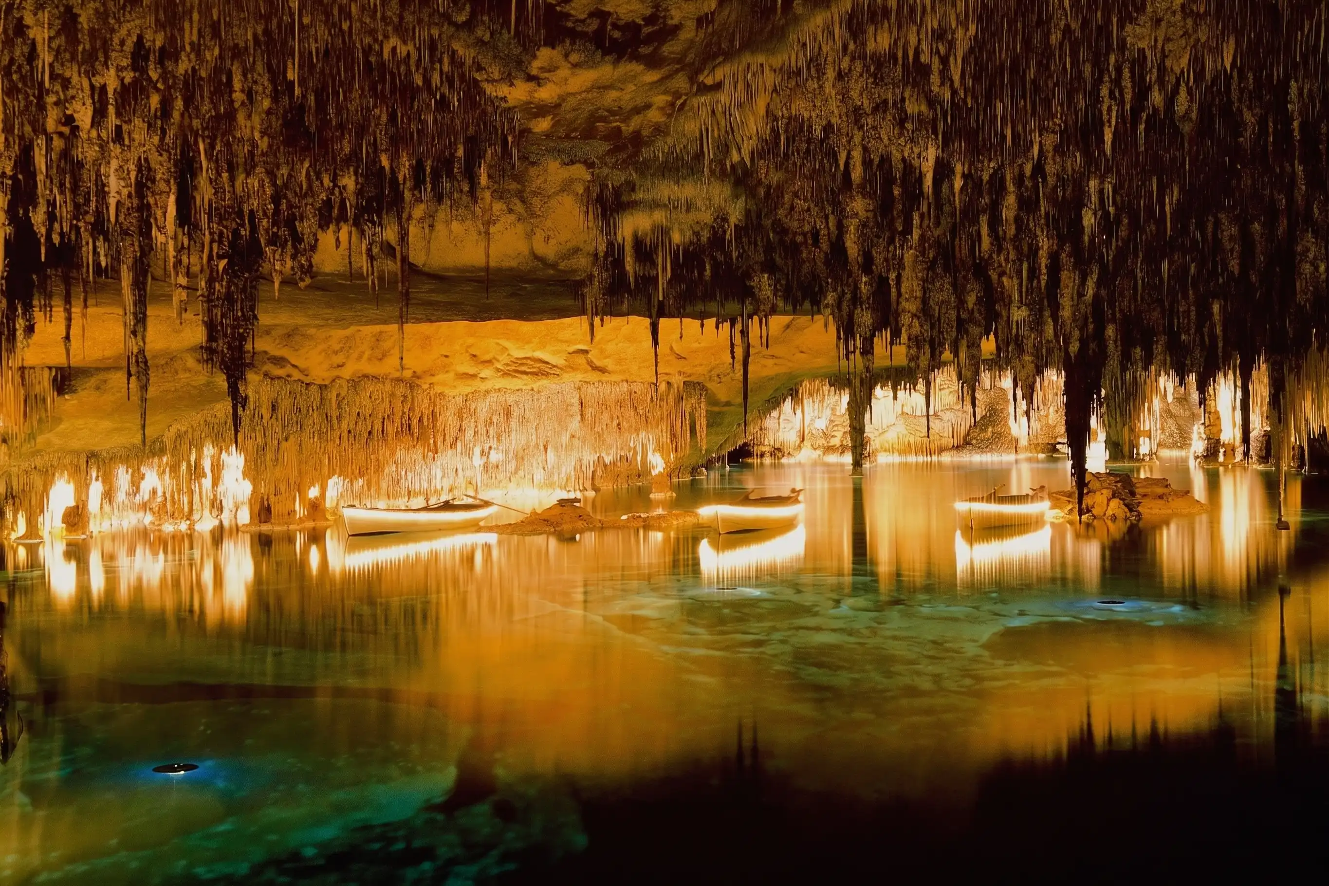 Small boats float on an underground lake inside a large cave illuminated by warm golden lights and stalactites.