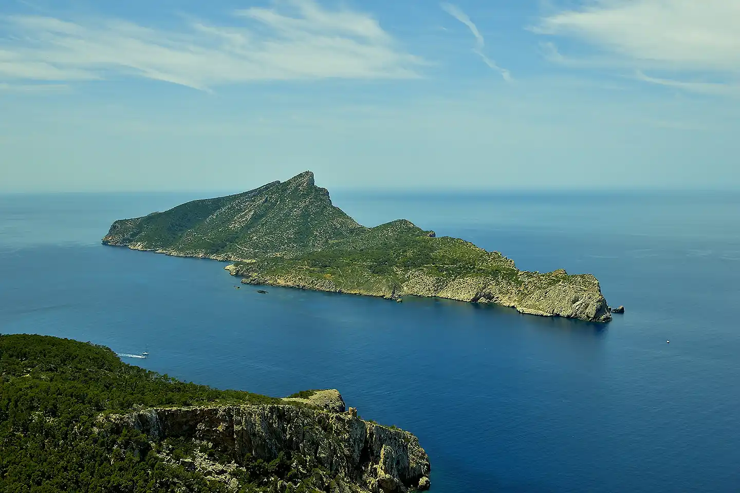 An elongated, rocky and green island sits in the middle of a calm blue sea under a partly cloudy sky.