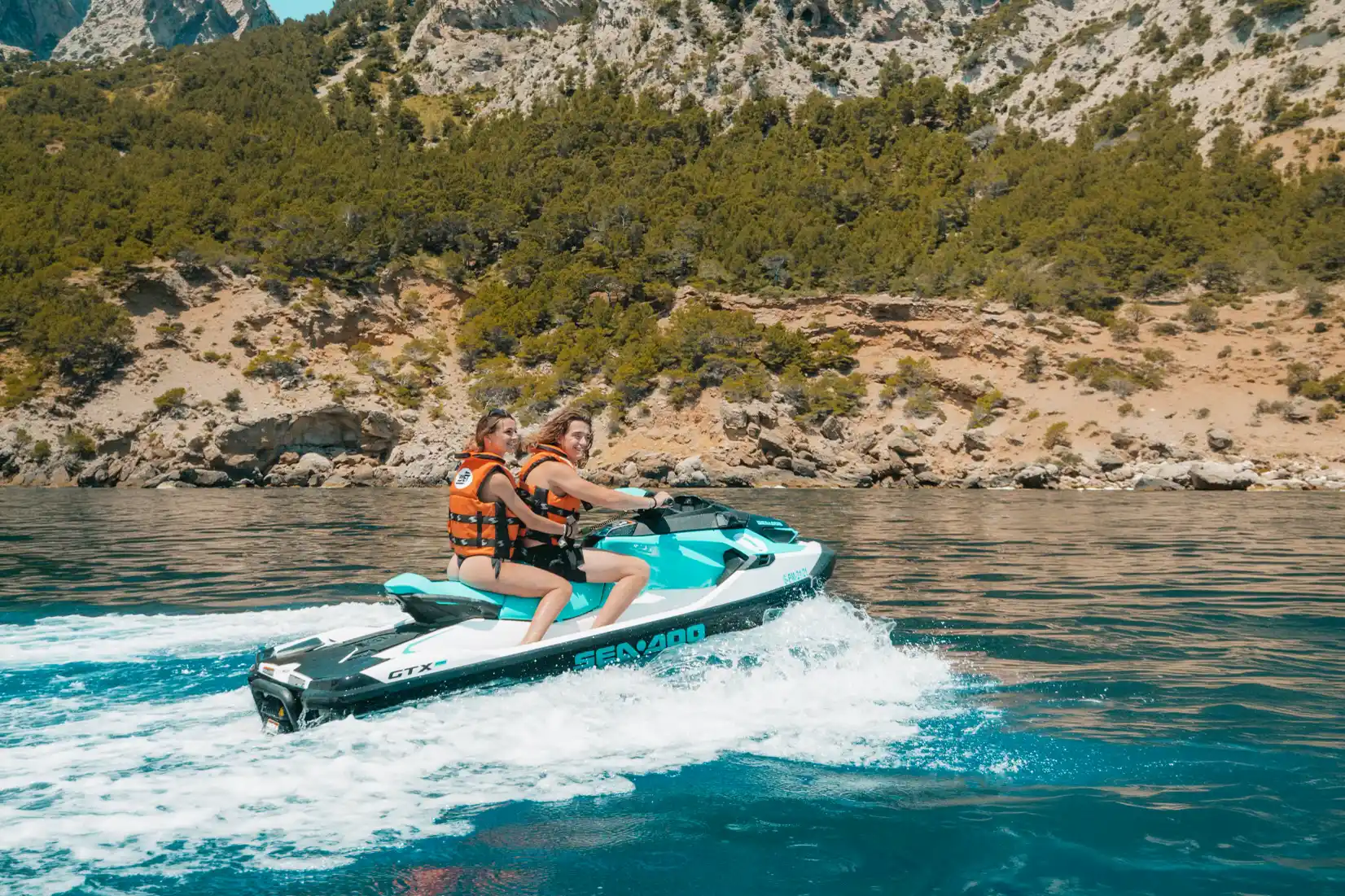 Two women in life jackets riding a fast jet ski on the ocean with a rocky, tree-covered coastline behind.