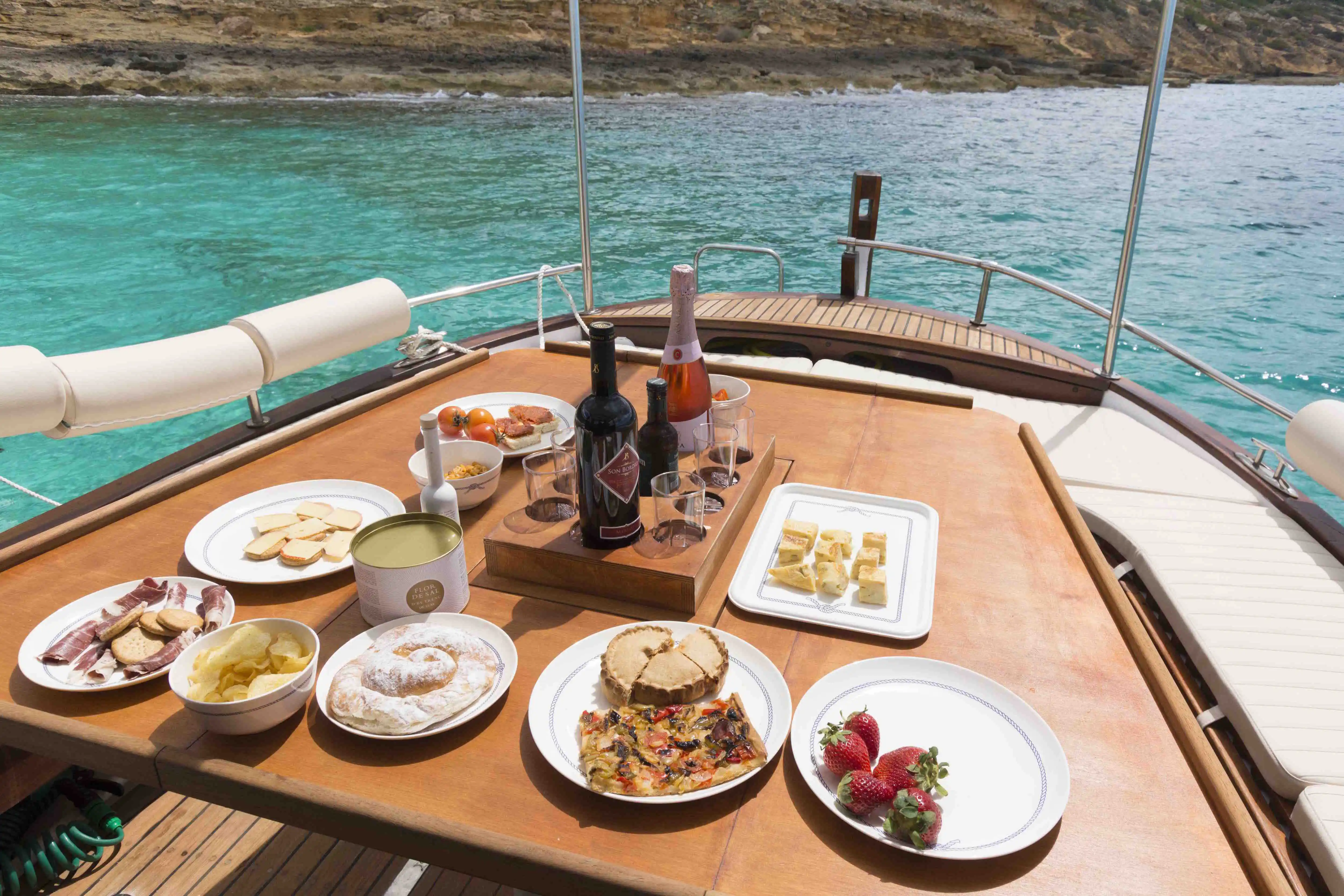 Ein Holztisch auf einem Bootsdeck voller Essen, Snacks und Weinflaschen auf dem türkisfarbenen Meer auf Mallorca. Ein Holztisch auf einem Bootsdeck voller Essen, Snacks und Weinflaschen auf dem türkisfarbenen Meer auf Mallorca.