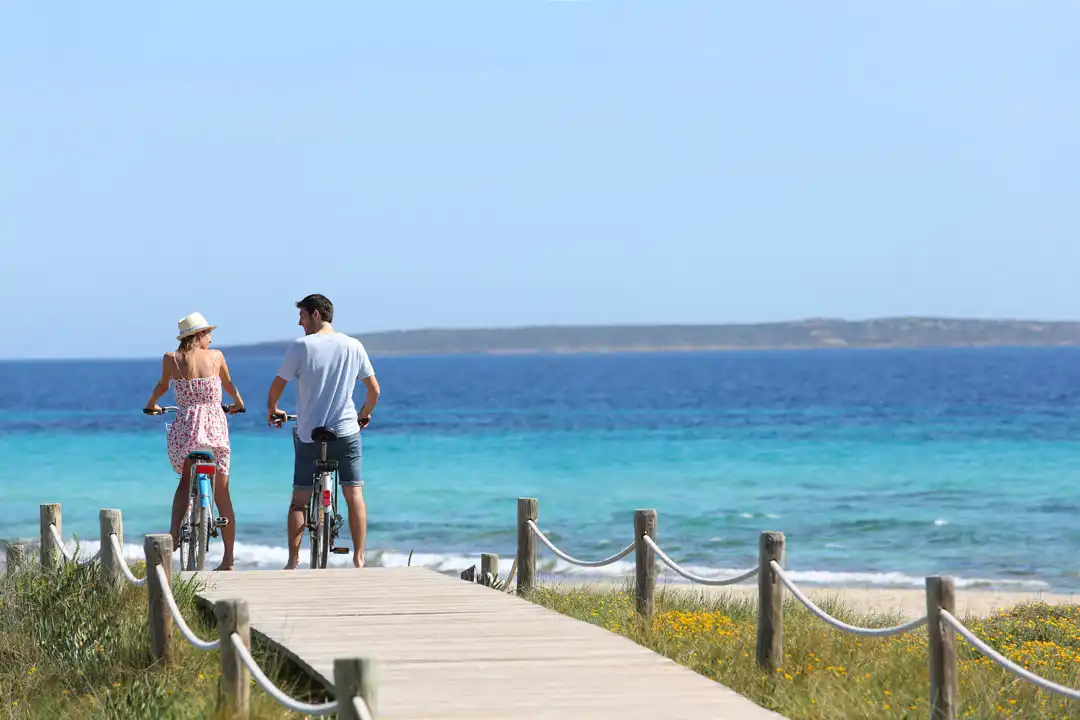 A couple riding bicycles stands on a wooden path looking out at the beautiful turquoise sea water.