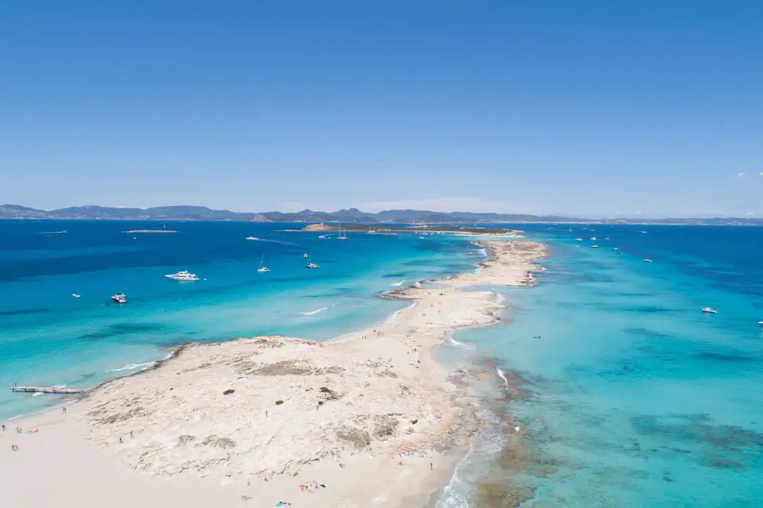 Aerial view of a narrow sandspit beach surrounded by turquoise and deep blue ocean waters under a clear sky.