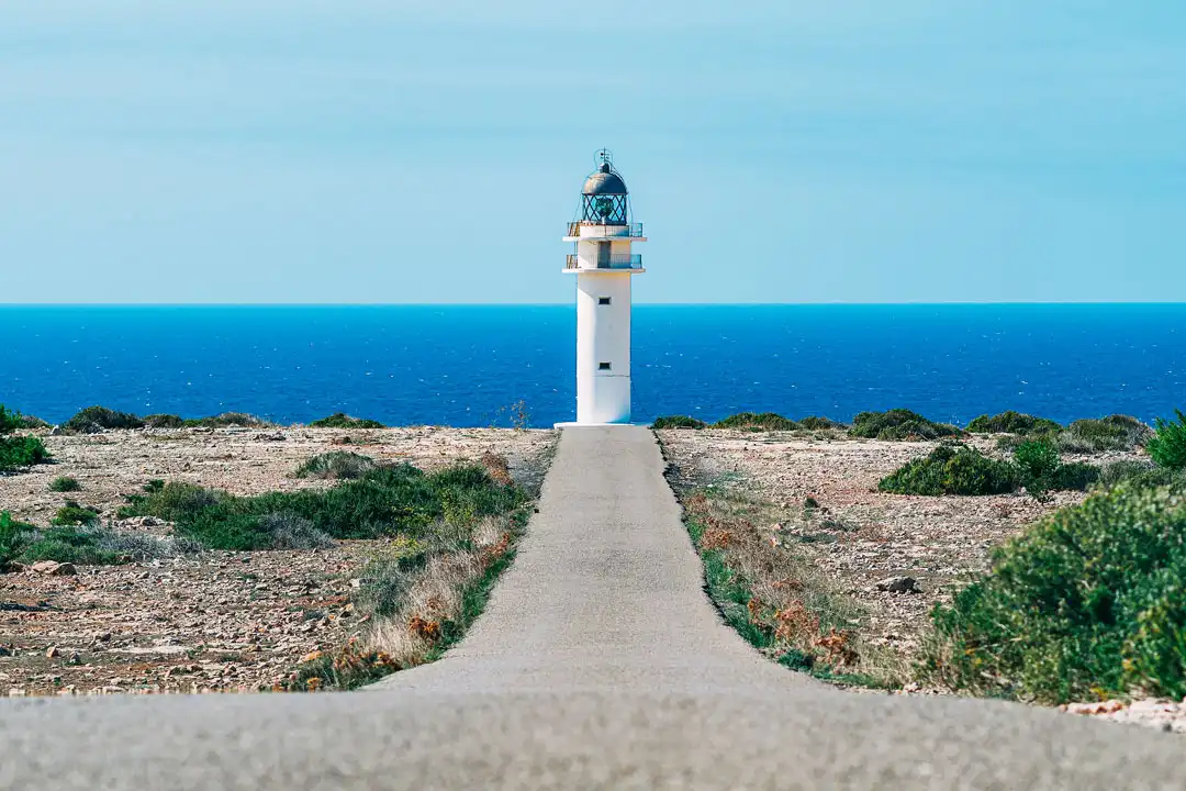 A straight asphalt road leads toward a white lighthouse standing on a rugged coastline by the blue sea.
