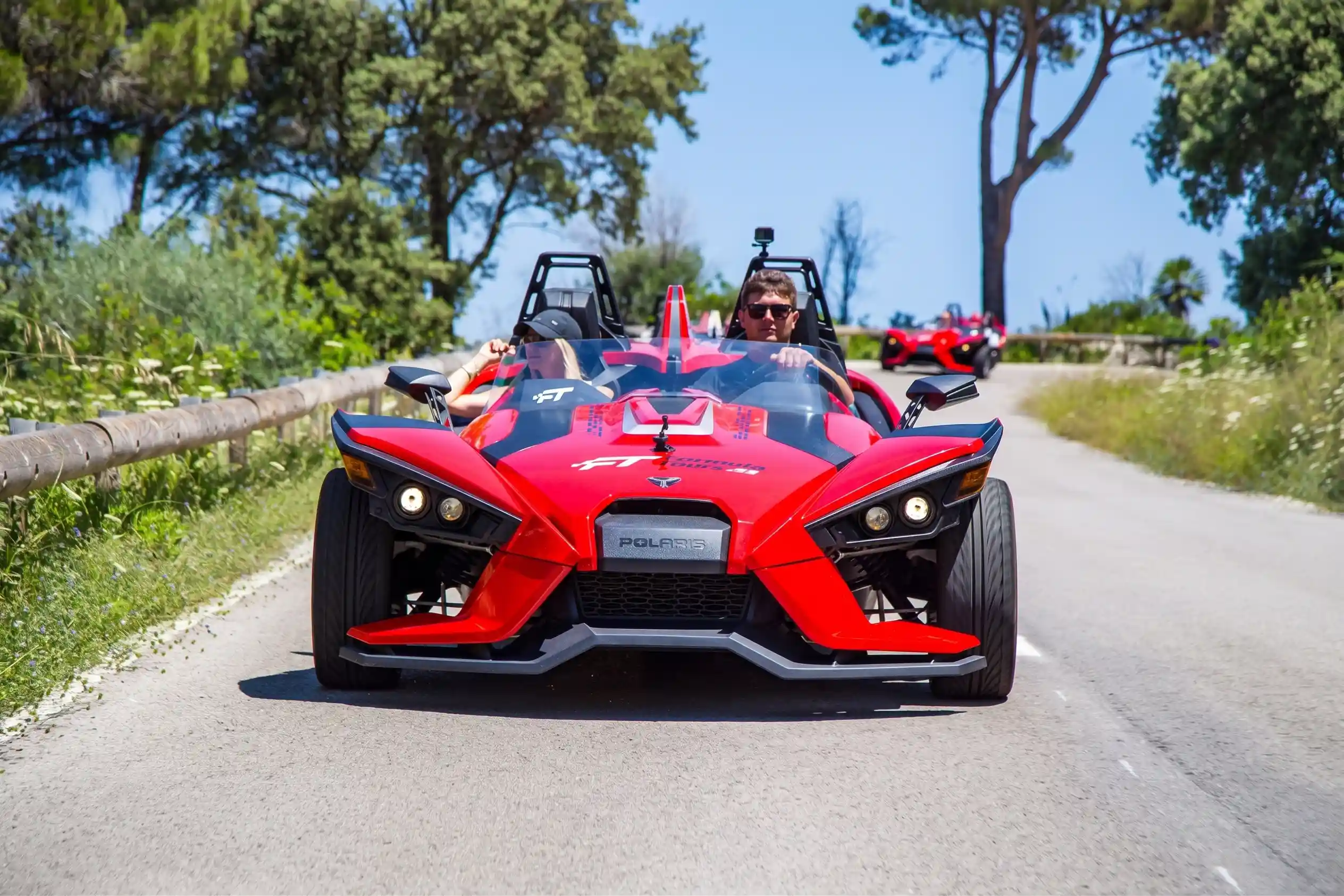 A man driving a red three-wheeled Slingshot car on a paved road surrounded by green trees.