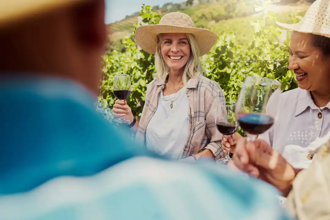 A group of smiling friends with straw hats toasts with glasses of red wine at an outdoor vineyard.
