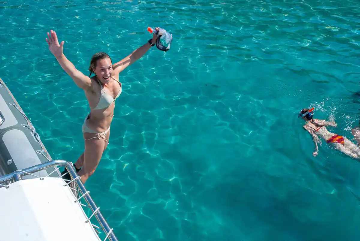 A happy woman jumping off a boat into clear turquoise water while holding a snorkel mask.