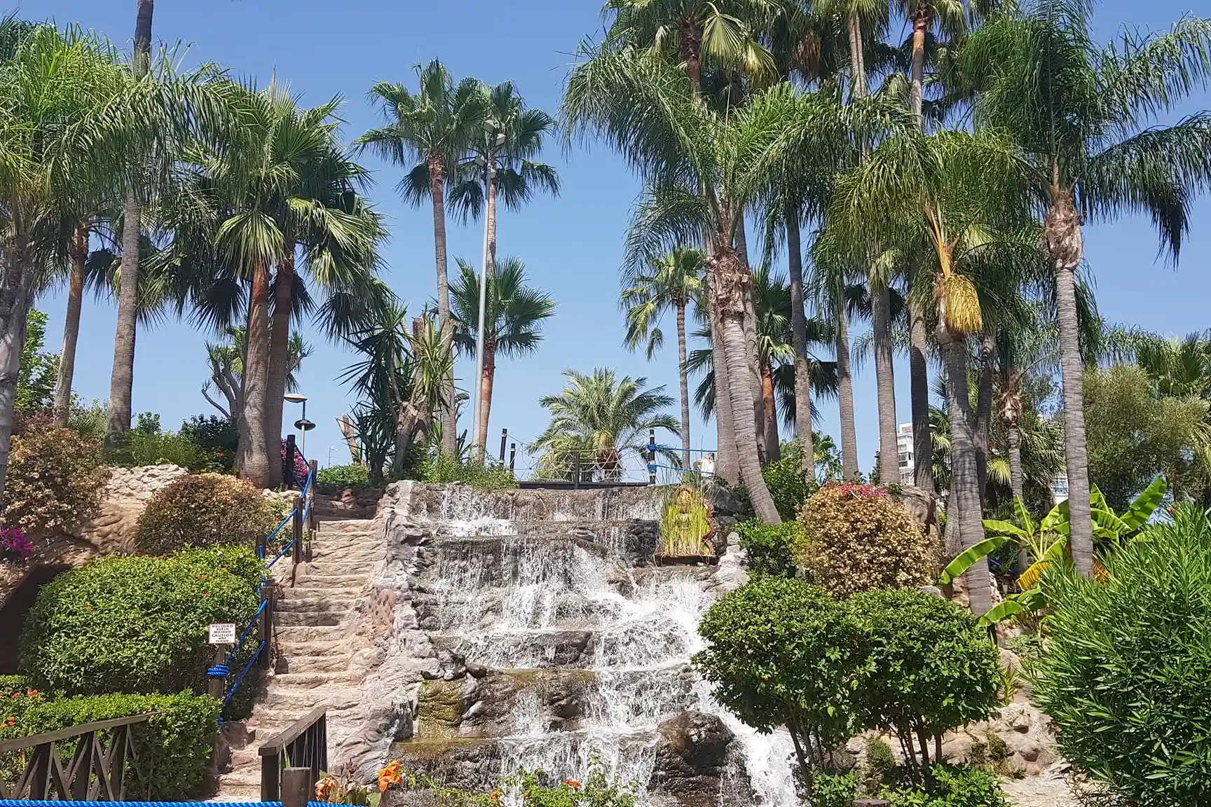 A stone waterfall at a mini-golf park surrounded by lush green bushes and tall palm trees.