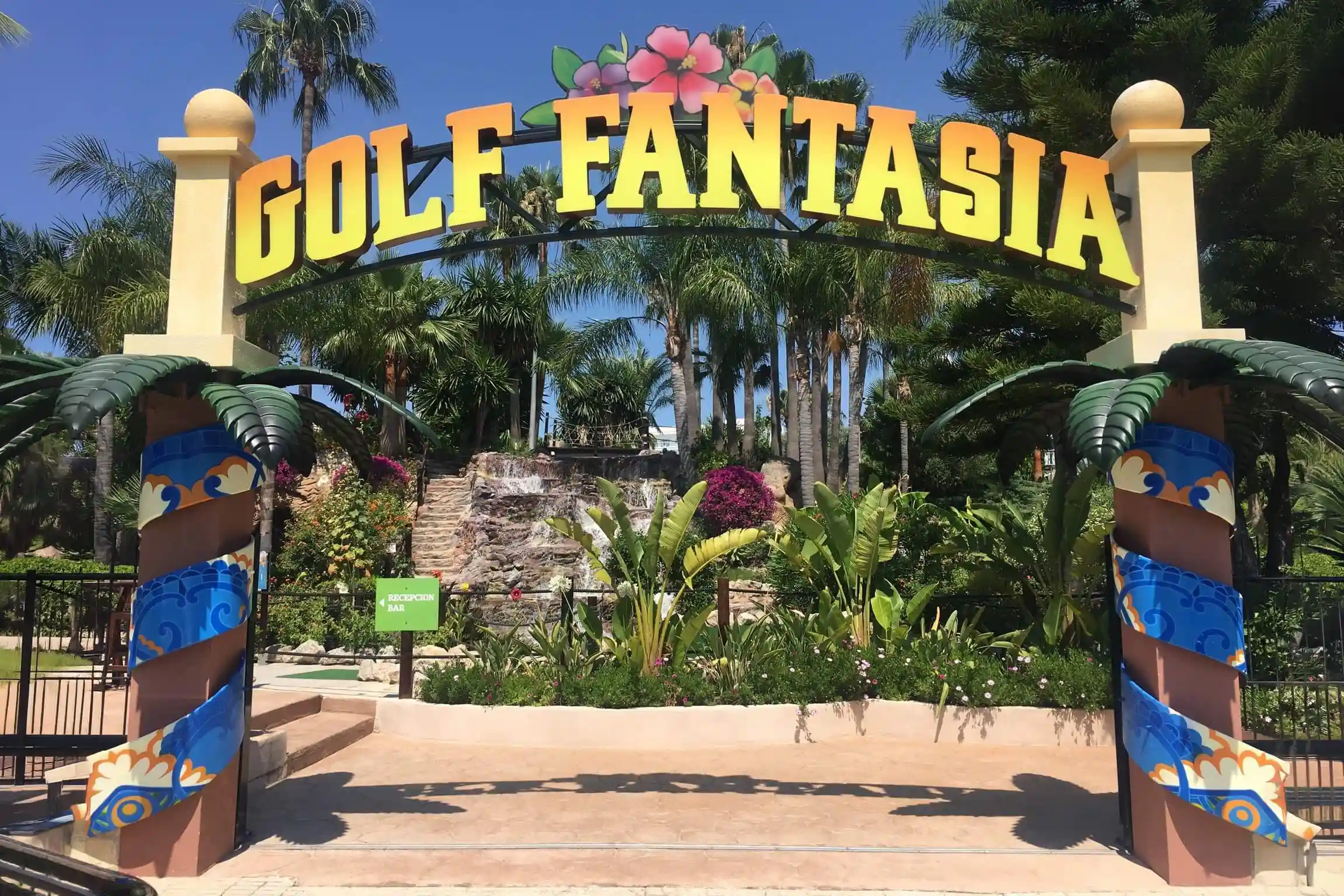 The colorful entrance arch of Golf Fantasia mini-golf park surrounded by tropical palm trees and plants.