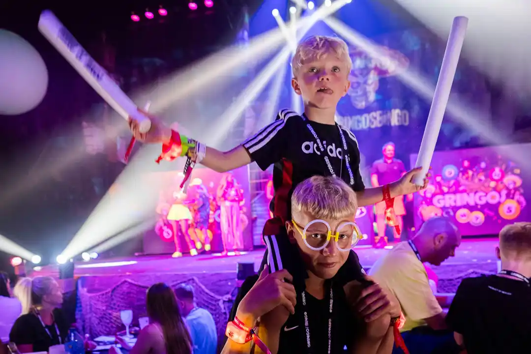 A young boy sits on another's shoulders, holding glowing foam sticks at a lively family rave event under spotlights.