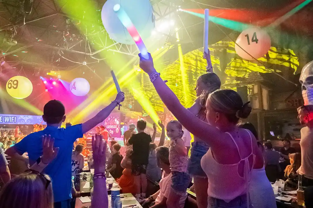 People wave glowing foam sticks at a lively rave party with large numbered balloons and bright lights.