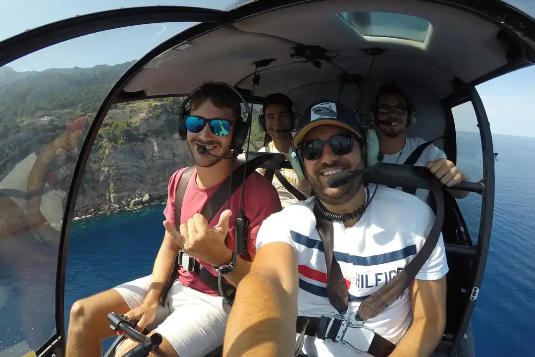 A group of friends takes a joyful selfie inside a helicopter cockpit flying over the blue sea and a rugged coast.