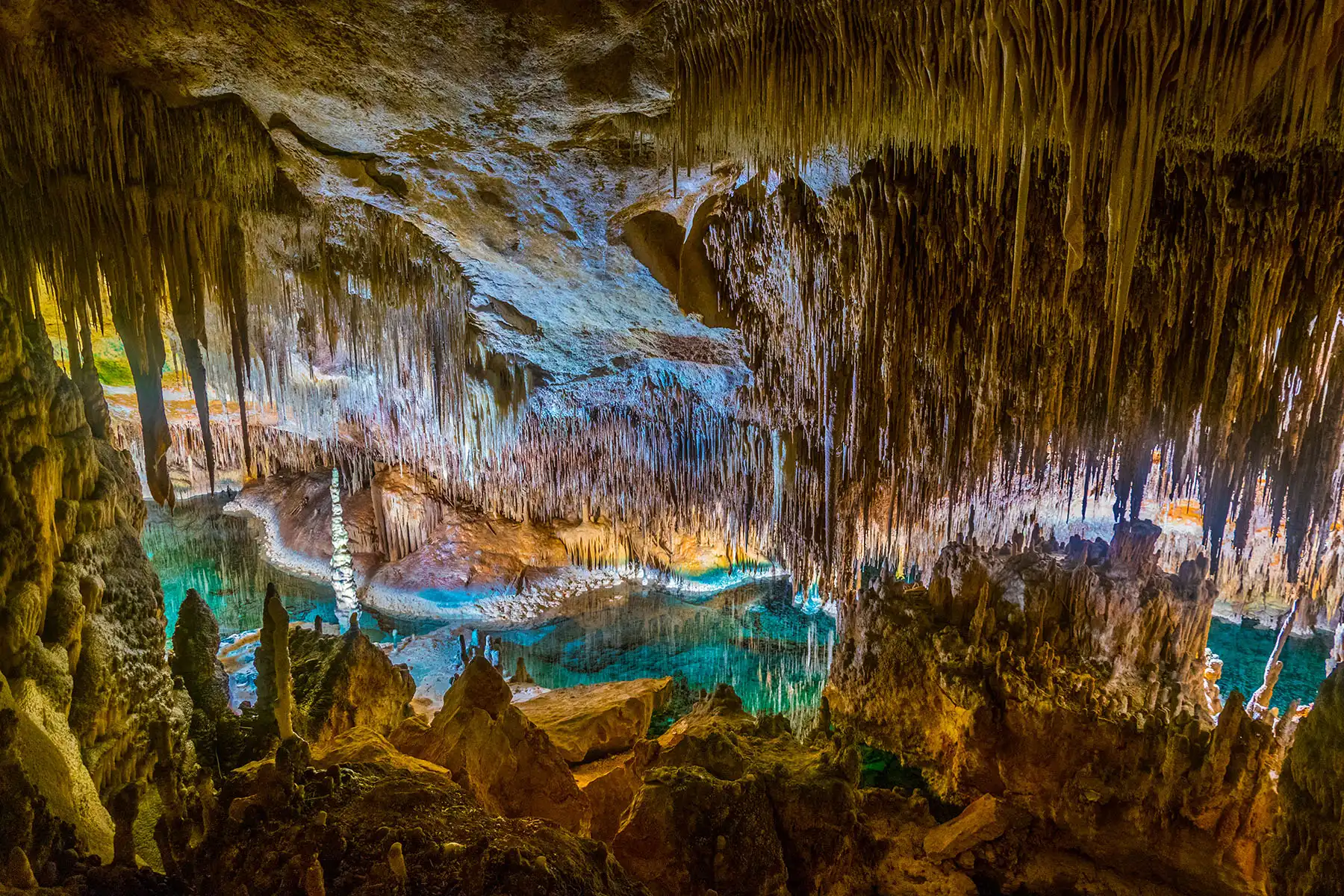 An impressive view of a turquoise underground lake illuminated inside a large cave filled with stalactites and stalagmites.