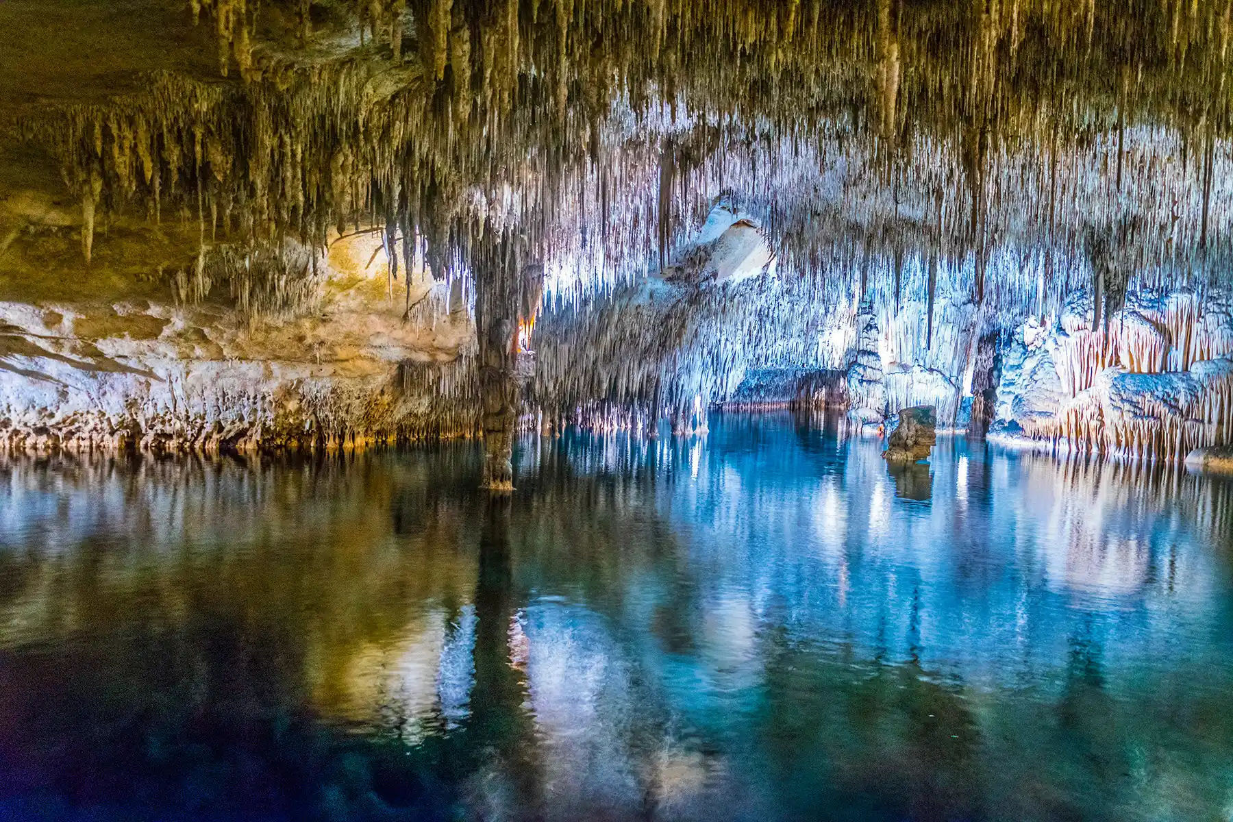 An impressive view of a turquoise underground lake illuminated inside a large cave filled with stalactites and stalagmites.