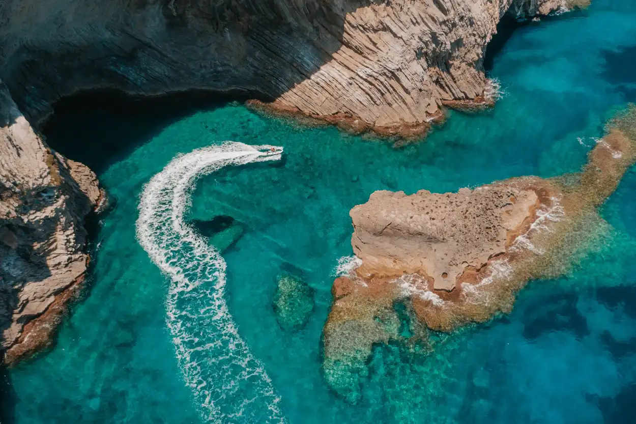 An aerial view of a jet ski making a white wake in turquoise ocean water near rocky cliffs and a sea cave. An aerial view of a jet ski making a white wake in turquoise ocean water near rocky cliffs and a sea cave.
