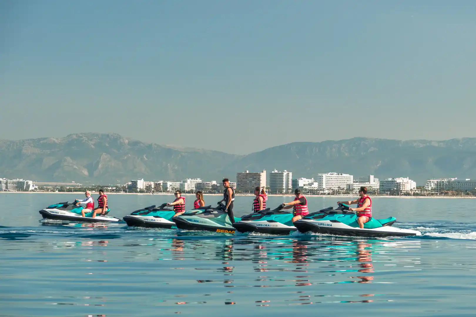 A group of people on turquoise jet skis lined up on the calm sea with buildings and mountains in Mallorca.